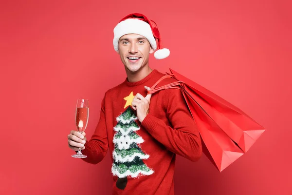 Homme souriant en santa chapeau tenant des sacs à provisions et une coupe de champagne sur fond rouge — Photo de stock