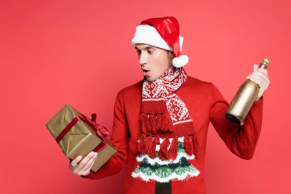 Excited man in santa hat holding bottle of champagne and gift box on red background — Stock Photo