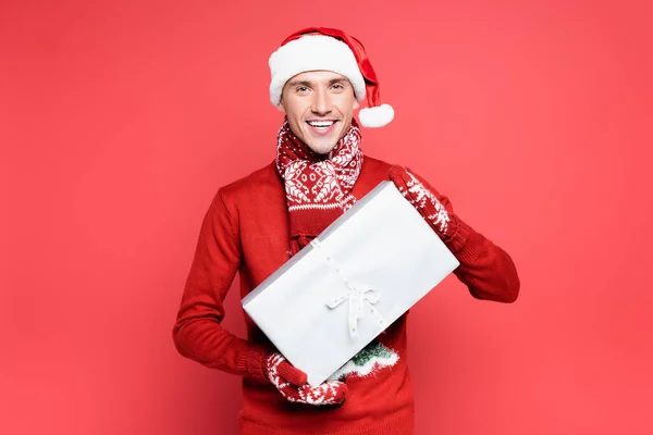 Smiling man in mittens and santa hat holding gift box on red background — Stock Photo