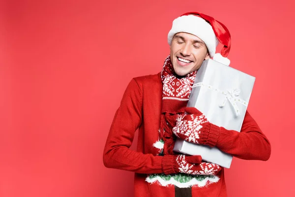 Lächelnder Mann mit Weihnachtsmütze und Fäustlingen, die Geschenk auf rotem Hintergrund halten — Stockfoto