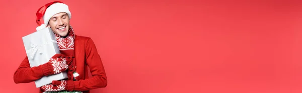 Sorrindo homem em mitenes e santa chapéu segurando caixa de presente isolado em vermelho, banner — Fotografia de Stock