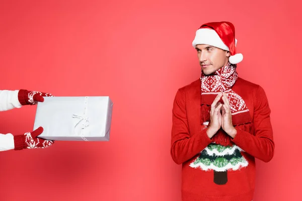 Schlauer Mann mit Schal und Weihnachtsmütze schaut auf Geschenkbox auf rotem Hintergrund — Stockfoto