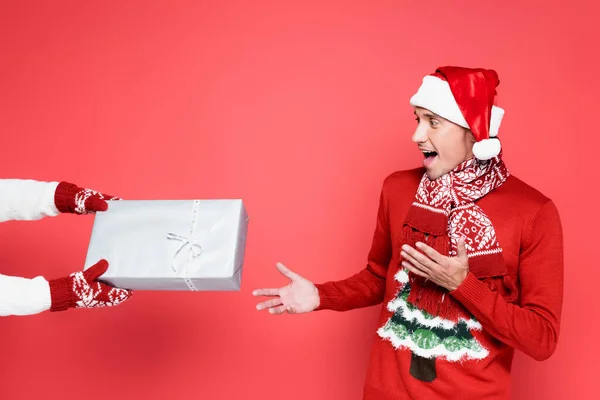 Шокований чоловік в капелюсі Санта і светр, дивлячись на присутність на червоному тлі — стокове фото