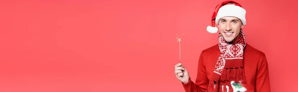 Giovane in cappello di Babbo Natale sorridente alla macchina fotografica mentre tiene sparkler isolato sul rosso, striscione — Foto stock