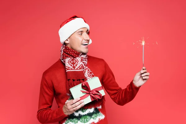 Smiling man in scarf and santa hat holding sparkler and gift box isolated on red — Stock Photo