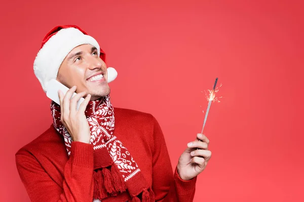 Uomo sorridente in cappello da Babbo Natale che tiene la scintilla mentre parla su smartphone isolato su rosso — Foto stock