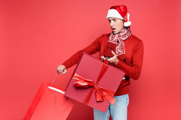 Young man in santa hat holding shopping bag and present on red background — Stock Photo