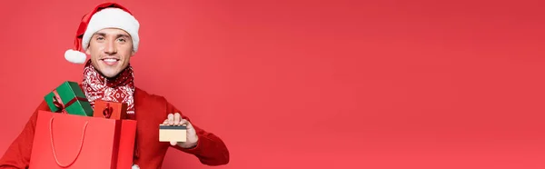 Hombre sonriente en sombrero de santa celebración de tarjeta de crédito y bolsa de compras con regalos aislados en rojo, pancarta - foto de stock