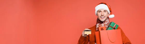 Uomo sorridente in cappello di Babbo Natale con carta di credito e shopping bag con scatole regalo isolate su rosso, banner — Foto stock