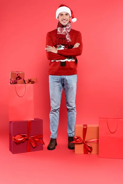 Hombre sonriente en sombrero de santa con brazos cruzados mirando a la cámara cerca de cajas de regalo y bolsas de compras sobre fondo rojo - foto de stock