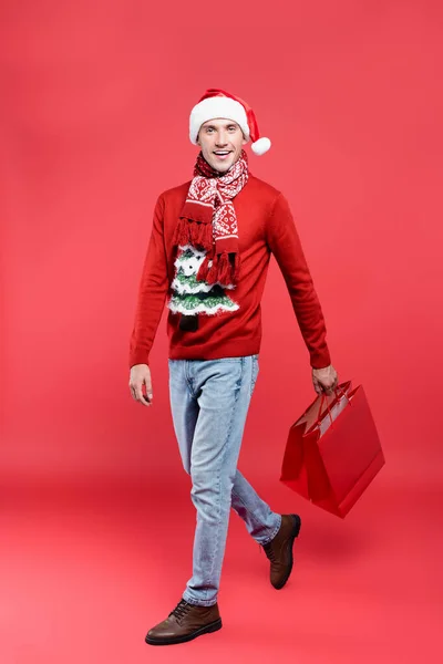 Homme souriant en santa chapeau tenant des sacs à provisions tout en marchant sur fond rouge — Photo de stock