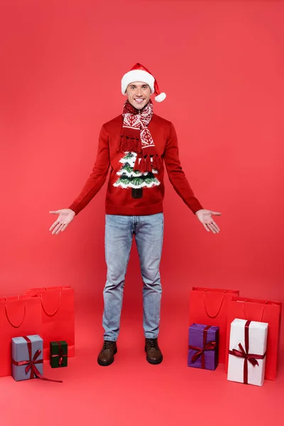 Sorrindo homem em santa chapéu apontando com as mãos em sacos de compras e caixas de presente no fundo vermelho — Fotografia de Stock