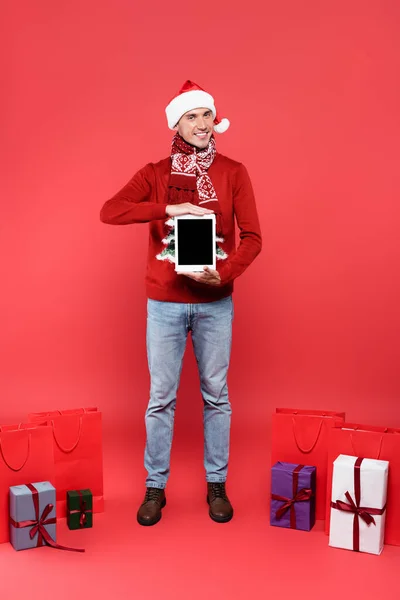 Hombre sonriente en sombrero de santa celebración tableta digital con pantalla en blanco cerca de regalos y bolsas de compras sobre fondo rojo - foto de stock