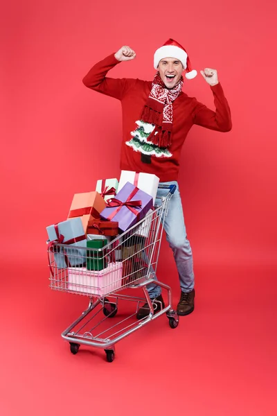 Hombre alegre en sombrero de santa mostrando sí gesto cerca de regalos en el carrito de la compra sobre fondo rojo - foto de stock