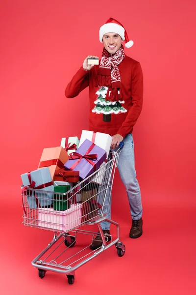 Lächelnder Mann mit Weihnachtsmütze und Kreditkarte neben Einkaufswagen mit Geschenken auf rotem Hintergrund — Stockfoto