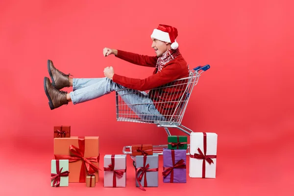 Seitenansicht eines jungen Mannes mit Weihnachtsmütze, der in einem Einkaufswagen in der Nähe von Geschenkboxen auf rotem Hintergrund sitzt — Stockfoto
