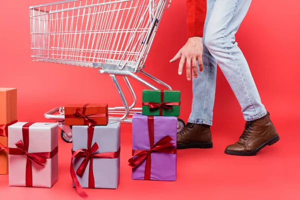 Vista recortada del hombre tirando de la mano a las cajas de regalo cerca del carrito de la compra sobre fondo rojo - foto de stock