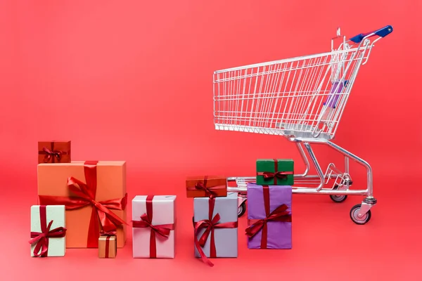 Gift boxes with ribbons near shopping cart on red background — Stock Photo