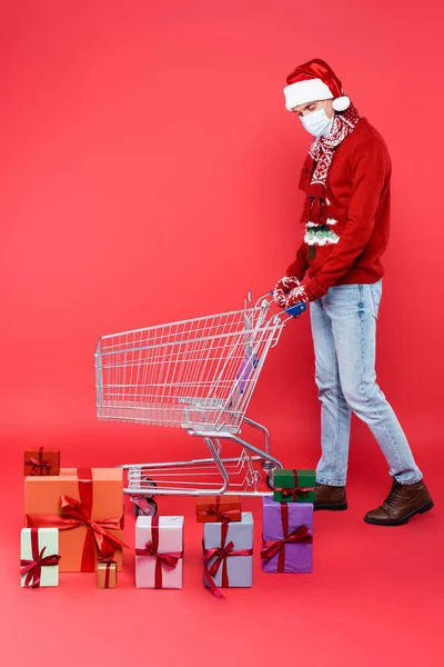 Homme en santa chapeau et masque médical regardant les cadeaux près du panier sur fond rouge — Photo de stock