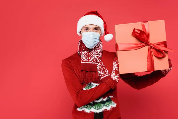 Человек в шляпе Санты, рукавицах и медицинской маске держит подарок с луком на красном фоне — стоковое фото