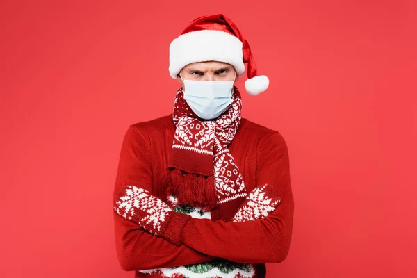 Homem irritado em máscara médica e chapéu de Papai Noel olhando para câmera isolada no vermelho — Fotografia de Stock