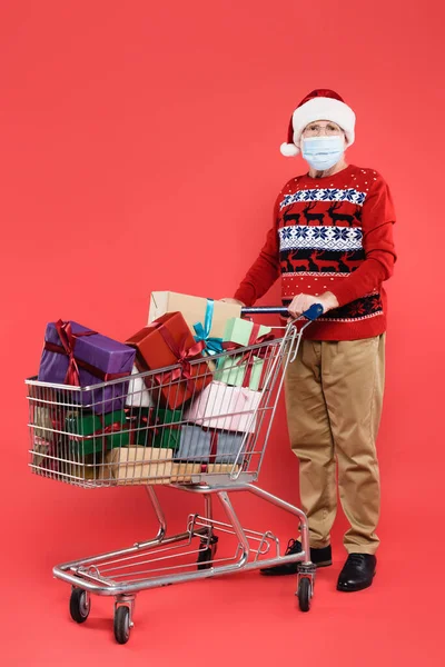 Uomo anziano in cappello di Babbo Natale e maschera medica in piedi vicino al carrello della spesa con regali su sfondo rosso — Foto stock