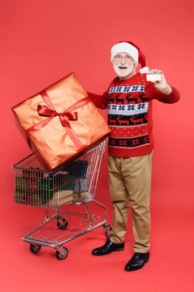 Cheerful senior man in santa hat showing credit card near shopping cart with presents on red background — Stock Photo