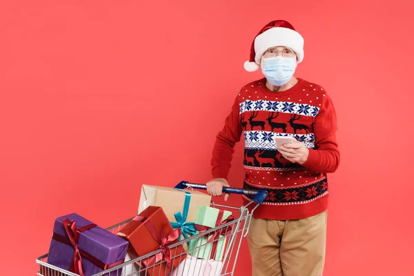 Senior man in santa hat and medical mask holding smartphone near cart with gift boxes isolated on red — Stock Photo