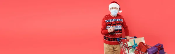 Senior man in santa hat and medical mask holding smartphone near shopping cart with gifts isolated on red, banner — Stock Photo
