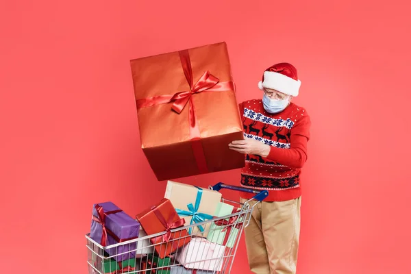 Homme âgé en santa chapeau et masque médical tenant cadeau près du panier avec des cadeaux isolés sur rouge — Photo de stock