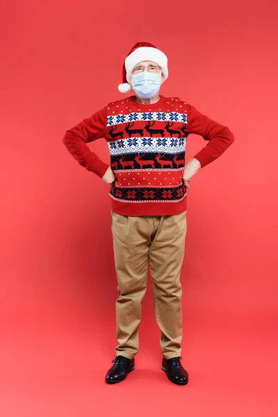 Homme âgé en santa chapeau et masque médical debout avec les mains sur les hanches sur fond rouge — Photo de stock