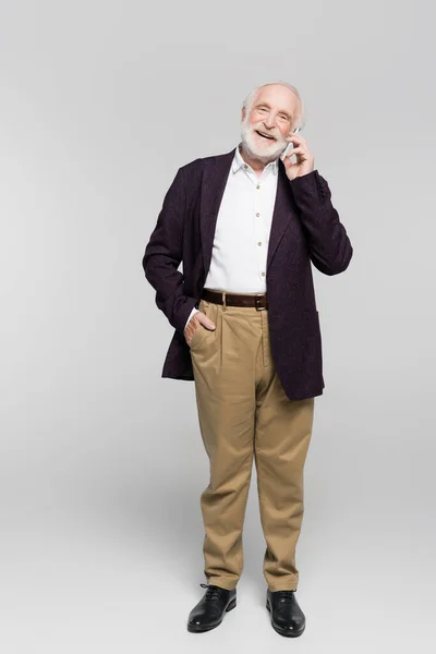 Hombre viejo sonriente con la mano en el bolsillo de los pantalones hablando en el teléfono inteligente sobre fondo gris - foto de stock