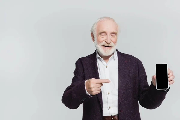 Hombre mayor sonriente apuntando con el dedo al teléfono inteligente con pantalla en blanco aislado en gris - foto de stock