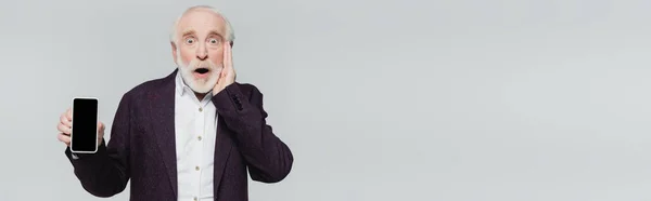 Hombre mayor sorprendido sosteniendo teléfono inteligente con pantalla en blanco aislado en gris, bandera - foto de stock