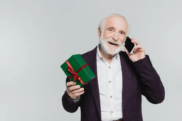 Hombre mayor sonriente sosteniendo regalo mientras habla en un teléfono inteligente aislado en gris - foto de stock