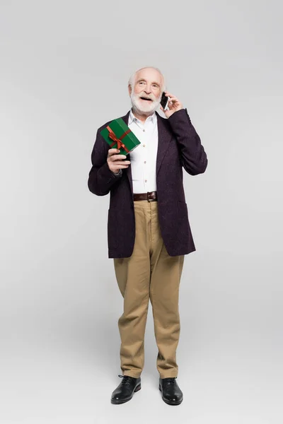 Hombre senior positivo hablando en el teléfono inteligente mientras sostiene la caja de regalo sobre fondo gris - foto de stock