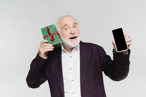 Aufgeregter Senior hält Smartphone mit leerem Bildschirm und Geschenk isoliert auf grau — Stockfoto