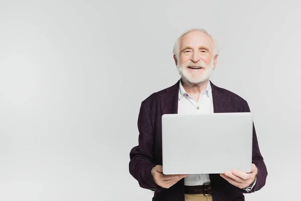 Smiling elderly man looking at camera while holding laptop isolated on grey — Stock Photo