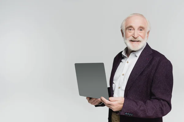 Hombre de pelo gris en camisa y chaqueta sonriendo a la cámara mientras sostiene el ordenador portátil aislado en gris - foto de stock