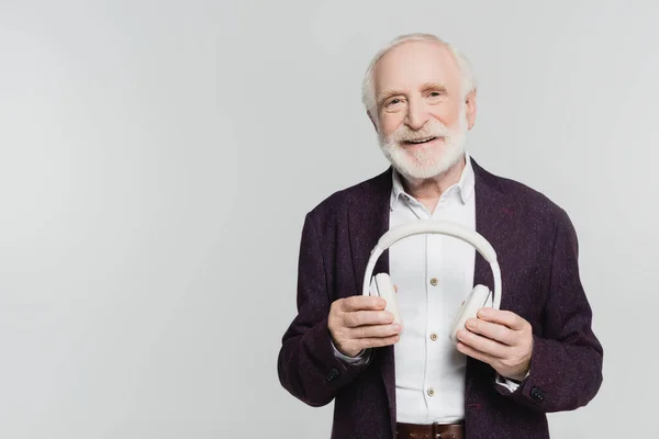 Hombre mayor sonriendo a la cámara mientras sostiene los auriculares aislados en gris - foto de stock