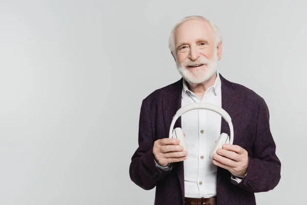 Homme âgé souriant à la caméra tout en tenant des écouteurs isolés sur gris — Photo de stock