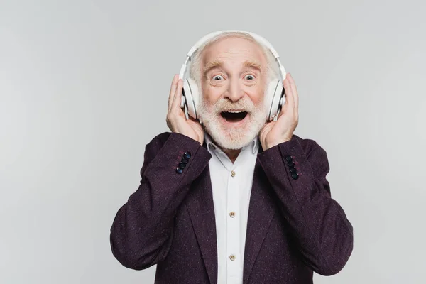 Hombre mayor emocionado mirando a la cámara mientras usa auriculares aislados en gris - foto de stock