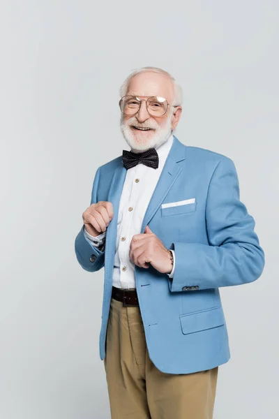 Smiling elderly man in eyeglasses and bow tie looking at camera isolated on grey — Stock Photo