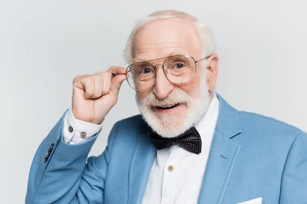 Positive senior man in blue jacket and bow tie touching eyeglasses isolated on grey — Stock Photo