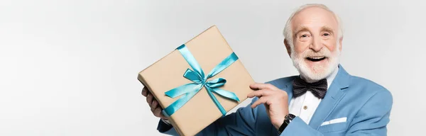 Homem idoso em gravata borboleta e casaco segurando caixa de presente e sorrindo para a câmera isolada em cinza, banner — Fotografia de Stock
