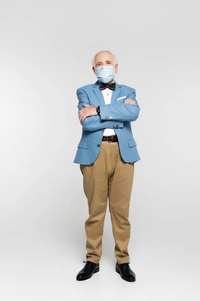 Uomo anziano in maschera medica in piedi con le braccia incrociate su sfondo grigio — Foto stock