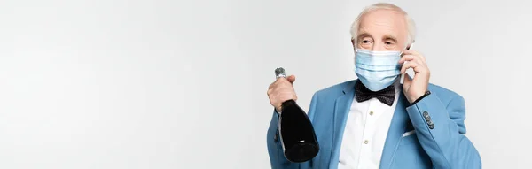 Старший чоловік в медичній масці розмовляє на смартфоні і тримає пляшку шампанського ізольовано на сірому, банер — стокове фото