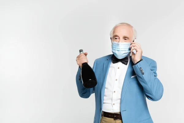 Elderly man in medical mask and bow tie holding bottle of champagne while talking on smartphone isolated on grey — Stock Photo