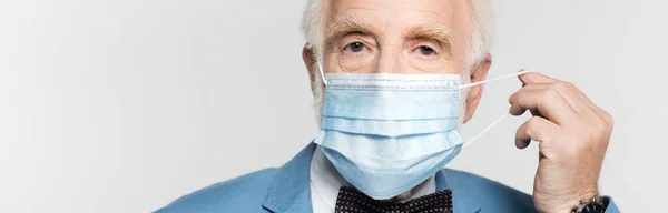 Elderly man holding medical mask isolated on grey, banner — Stock Photo