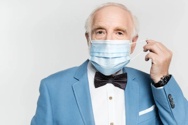 Elderly man in bow tie and jacket holding medical mask isolated on grey — Stock Photo